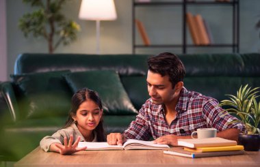 Bir ders kitabından kızı eğitmek, Hintli genç bir baba, modern evdeki masada birlikte otururken çocuk eğitimine yardım etmek, Asyalı bir baba, ev ödevi, aile bağı ve eğitimi olan bir kıza rehberlik etmek.