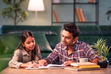 Bir ders kitabından kızı eğitmek, Hintli genç bir baba, modern evdeki masada birlikte otururken çocuk eğitimine yardım etmek, Asyalı bir baba, ev ödevi, aile bağı ve eğitimi olan bir kıza rehberlik etmek.