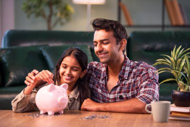 Kızına kumbarada para biriktirmeyi öğretiyor, Hintli baba çocuğuna mali birikimleri öğretiyor, modern evdeki masada birlikte oturuyor, Asyalı baba kızla bağ kuruyor ve para yönetimi öğretiyor.