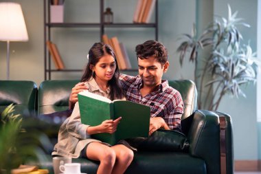 Birlikte kitap okuyoruz, Hintli baba ve kızı modern evdeki koltukta oturuyor Asyalı baba genç kızla bağ kuruyor, aile kalitesi, ev içi öğrenme ve duygusal bağ paylaşıyor.