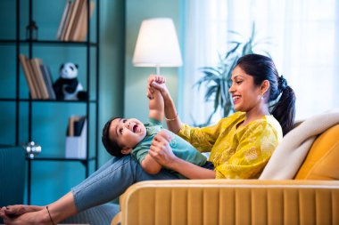 Hintli genç anne, modern şık ev iç mekanında rahat bir kanepede bebeğe sarılıyor sıcaklığını, şefkatini ve duygusal aşkını ifade ederken güzel anneliğin tadını çıkarıyor.