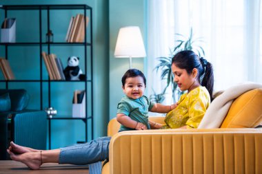 Hintli genç anne, modern şık ev iç mekanında rahat bir kanepede bebeğe sarılıyor sıcaklığını, şefkatini ve duygusal aşkını ifade ederken güzel anneliğin tadını çıkarıyor.