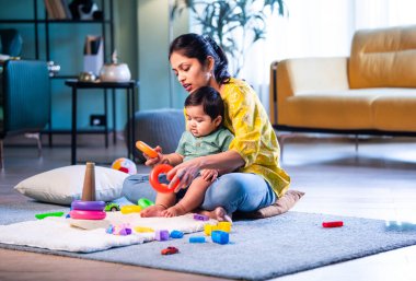 Hintli çocuk modern oturma odasında halıda otururken genç anneyle yüzük oyuncakları oynuyor. Sevgi, şefkat, bağ ve ebeveynliğin mutluluğunu paylaşıyor.