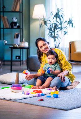 Hintli çocuk modern oturma odasında halıda otururken genç anneyle yüzük oyuncakları oynuyor. Sevgi, şefkat, bağ ve ebeveynliğin mutluluğunu paylaşıyor.