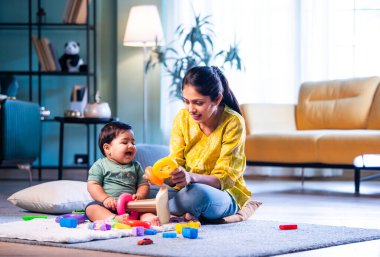 Hintli çocuk modern oturma odasında halıda otururken genç anneyle yüzük oyuncakları oynuyor. Sevgi, şefkat, bağ ve ebeveynliğin mutluluğunu paylaşıyor.