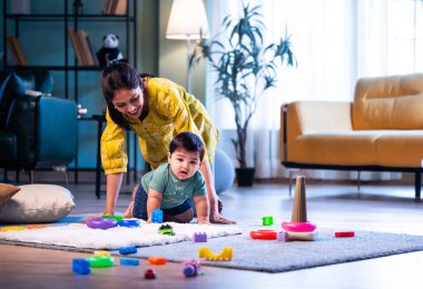 Hintli çocuk halıda genç annesiyle birlikte modern oturma odasında sürünüyor, neşeli anları paylaşıyor, sevgi, şefkat, bağ kurma ve erken çocukluk gelişimini evde besliyor.