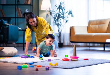 Hintli çocuk halıda genç annesiyle birlikte modern oturma odasında sürünüyor, neşeli anları paylaşıyor, sevgi, şefkat, bağ kurma ve erken çocukluk gelişimini evde besliyor.