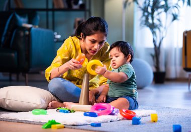 Hintli çocuk modern oturma odasında halıda otururken genç anneyle yüzük oyuncakları oynuyor. Sevgi, şefkat, bağ ve ebeveynliğin mutluluğunu paylaşıyor.