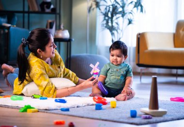 Hintli çocuk modern oturma odasında halıda otururken genç anneyle yüzük oyuncakları oynuyor. Sevgi, şefkat, bağ ve ebeveynliğin mutluluğunu paylaşıyor.