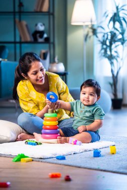Hintli çocuk modern oturma odasında halıda otururken genç anneyle yüzük oyuncakları oynuyor. Sevgi, şefkat, bağ ve ebeveynliğin mutluluğunu paylaşıyor.