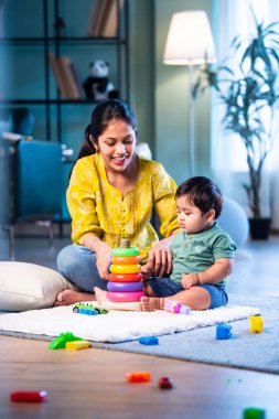 Hintli çocuk modern oturma odasında halıda otururken genç anneyle yüzük oyuncakları oynuyor. Sevgi, şefkat, bağ ve ebeveynliğin mutluluğunu paylaşıyor.