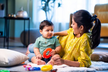 Hintli çocuk modern oturma odasında halıda otururken genç anneyle yüzük oyuncakları oynuyor. Sevgi, şefkat, bağ ve ebeveynliğin mutluluğunu paylaşıyor.