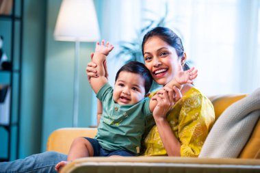 Hintli genç anne, modern şık ev iç mekanında rahat bir kanepede bebeğe sarılıyor sıcaklığını, şefkatini ve duygusal aşkını ifade ederken güzel anneliğin tadını çıkarıyor.