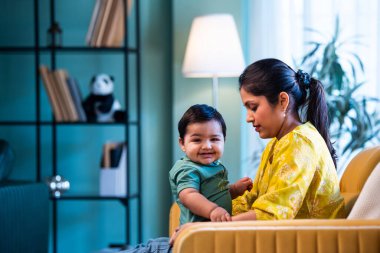 Hintli genç anne, modern şık ev iç mekanında rahat bir kanepede bebeğe sarılıyor sıcaklığını, şefkatini ve duygusal aşkını ifade ederken güzel anneliğin tadını çıkarıyor.