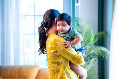 Hintli bir bebek annesinin omzunda uyur. Sıcak, modern bir evde sevgi dolu bir şekilde bebeğine sarılır ve onu rahatlatır. Huzurlu bir bağ, annelik, şefkat ve erken ebeveynlik gösterir.