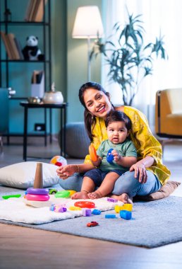 Hintli çocuk modern oturma odasında halıda otururken genç anneyle yüzük oyuncakları oynuyor. Sevgi, şefkat, bağ ve ebeveynliğin mutluluğunu paylaşıyor.