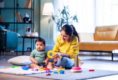 Hintli çocuk modern oturma odasında halıda otururken genç anneyle yüzük oyuncakları oynuyor. Sevgi, şefkat, bağ ve ebeveynliğin mutluluğunu paylaşıyor.