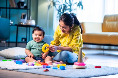 Hintli çocuk modern oturma odasında halıda otururken genç anneyle yüzük oyuncakları oynuyor. Sevgi, şefkat, bağ ve ebeveynliğin mutluluğunu paylaşıyor.