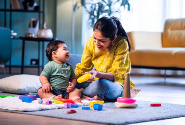 Hintli çocuk modern oturma odasında halıda otururken genç anneyle yüzük oyuncakları oynuyor. Sevgi, şefkat, bağ ve ebeveynliğin mutluluğunu paylaşıyor.