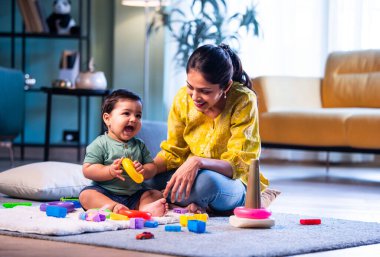 Hintli çocuk modern oturma odasında halıda otururken genç anneyle yüzük oyuncakları oynuyor. Sevgi, şefkat, bağ ve ebeveynliğin mutluluğunu paylaşıyor.