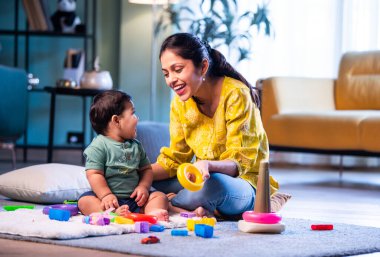 Hintli çocuk modern oturma odasında halıda otururken genç anneyle yüzük oyuncakları oynuyor. Sevgi, şefkat, bağ ve ebeveynliğin mutluluğunu paylaşıyor.