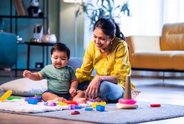Hintli çocuk modern oturma odasında halıda otururken genç anneyle yüzük oyuncakları oynuyor. Sevgi, şefkat, bağ ve ebeveynliğin mutluluğunu paylaşıyor.
