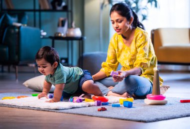 Hintli çocuk halıda genç annesiyle birlikte modern oturma odasında sürünüyor, neşeli anları paylaşıyor, sevgi, şefkat, bağ kurma ve erken çocukluk gelişimini evde besliyor.