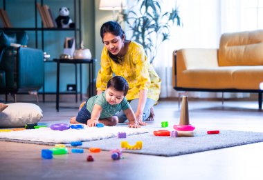 Hintli çocuk halıda genç annesiyle birlikte modern oturma odasında sürünüyor, neşeli anları paylaşıyor, sevgi, şefkat, bağ kurma ve erken çocukluk gelişimini evde besliyor.