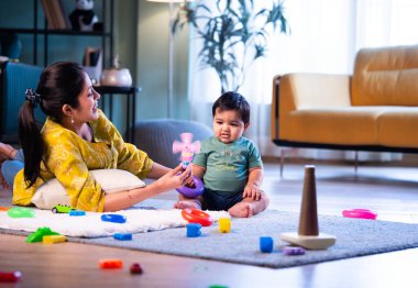 Hintli çocuk modern oturma odasında halıda otururken genç anneyle yüzük oyuncakları oynuyor. Sevgi, şefkat, bağ ve ebeveynliğin mutluluğunu paylaşıyor.