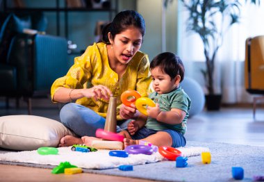 Hintli çocuk modern oturma odasında halıda otururken genç anneyle yüzük oyuncakları oynuyor. Sevgi, şefkat, bağ ve ebeveynliğin mutluluğunu paylaşıyor.