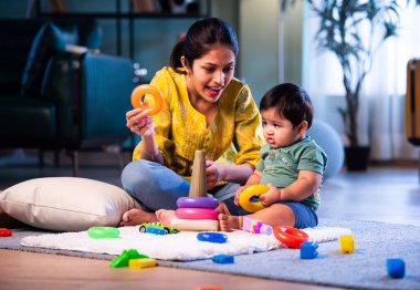 Hintli çocuk modern oturma odasında halıda otururken genç anneyle yüzük oyuncakları oynuyor. Sevgi, şefkat, bağ ve ebeveynliğin mutluluğunu paylaşıyor.