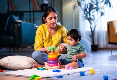 Hintli çocuk modern oturma odasında halıda otururken genç anneyle yüzük oyuncakları oynuyor. Sevgi, şefkat, bağ ve ebeveynliğin mutluluğunu paylaşıyor.