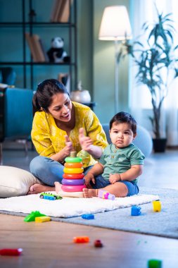 Hintli çocuk modern oturma odasında halıda otururken genç anneyle yüzük oyuncakları oynuyor. Sevgi, şefkat, bağ ve ebeveynliğin mutluluğunu paylaşıyor.