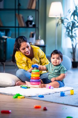 Hintli çocuk modern oturma odasında halıda otururken genç anneyle yüzük oyuncakları oynuyor. Sevgi, şefkat, bağ ve ebeveynliğin mutluluğunu paylaşıyor.