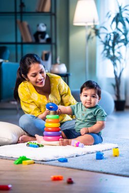 Hintli çocuk modern oturma odasında halıda otururken genç anneyle yüzük oyuncakları oynuyor. Sevgi, şefkat, bağ ve ebeveynliğin mutluluğunu paylaşıyor.