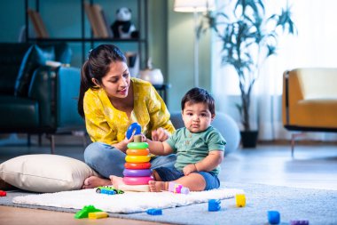 Hintli çocuk modern oturma odasında halıda otururken genç anneyle yüzük oyuncakları oynuyor. Sevgi, şefkat, bağ ve ebeveynliğin mutluluğunu paylaşıyor.