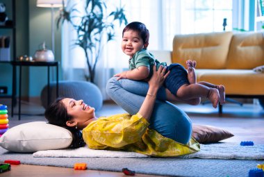 Hintli anne, bebeği ayaklarının üzerinde kaldırırken, halının üzerinde yatarken, eğlenceli oyunlar oynarken, bebek mutlu bir şekilde gülümserken, sevgi dolu anları paylaşırken, sevgi, şefkat ve neşe içinde modern ev ortamında