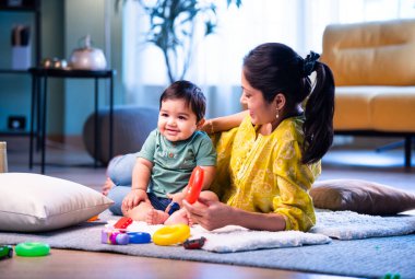 Hintli çocuk modern oturma odasında halıda otururken genç anneyle yüzük oyuncakları oynuyor. Sevgi, şefkat, bağ ve ebeveynliğin mutluluğunu paylaşıyor.