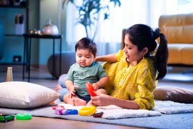 Hintli çocuk modern oturma odasında halıda otururken genç anneyle yüzük oyuncakları oynuyor. Sevgi, şefkat, bağ ve ebeveynliğin mutluluğunu paylaşıyor.