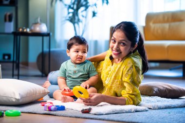 Hintli çocuk modern oturma odasında halıda otururken genç anneyle yüzük oyuncakları oynuyor. Sevgi, şefkat, bağ ve ebeveynliğin mutluluğunu paylaşıyor.