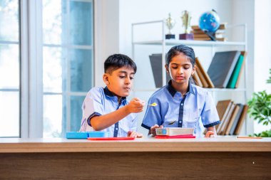 Sınıfta üniforma giyerek Tiffin yiyen ve paylaşan Hintli okul çocukları, okul günü boyunca arkadaşlarıyla keyifli ve samimi bir çocukluk anıyla öğle yemeği molasının tadını çıkarıyorlar.