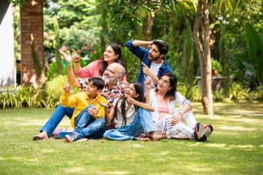 Hint çoklu nesiller ailesi yeşil çimlerin üzerinde birlikte oturan bir şeyi işaret ediyor ve altı mutlu üye bahçede oturmuş neşeli ifadeleri olan hayali bir ürün sergiliyor.