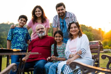 Hintli bir aile bankta oturmuş kameraya gülümsüyor, sıcak bağlanma anlarının tadını çıkarıyor, eğlenceli sohbetler yapıyor, neşeli kahkahalar atıyor ve açık havada rahat vakit geçiriyorlar.