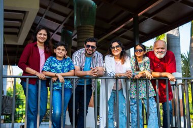 Korkuluklarda dikilen Hintli aile dışarıda gülümsüyor, birlikte eğleniyorlar, neşeli çoklu jenerasyon portresi, büyükanne ve büyükbabalar, çocuklar güpegündüz mutlu mesut kaynaşıyorlar.