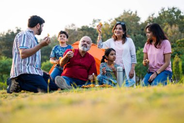 Hint ailesi dışarıda kamp yaparken abur cubur yiyor, kameraya bakıyor, neşeli bağlanma anlarının tadını çıkarıyor, kahkahaları paylaşıyor, gevşemiş sohbetler yapıyor, sıcak etkileşimler kuruyor ve birlikte keyifli güzel vakit geçiriyorlar.
