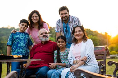 Hintli bir aile bankta oturmuş kameraya gülümsüyor, sıcak bağlanma anlarının tadını çıkarıyor, eğlenceli sohbetler yapıyor, neşeli kahkahalar atıyor ve açık havada rahat vakit geçiriyorlar.