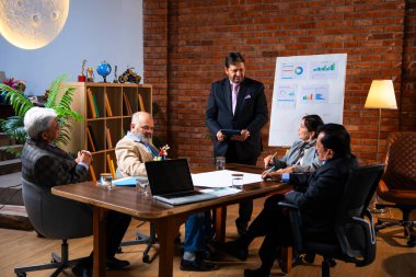 Hintli iş adamları projeyi tartışırken sunucu ayakta duruyor. Hintli iş adamı kıdemli yönetici ekibine stratejiyi açıklıyor.