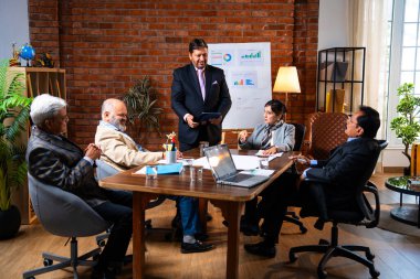 Hintli iş adamları projeyi tartışırken sunucu ayakta duruyor. Hintli iş adamı kıdemli yönetici ekibine stratejiyi açıklıyor.