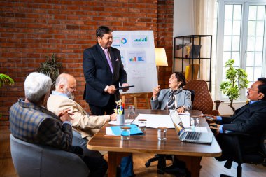 Hintli iş adamları projeyi tartışırken sunucu ayakta duruyor. Hintli iş adamı kıdemli yönetici ekibine stratejiyi açıklıyor.