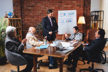 Hintli iş adamları projeyi tartışırken sunucu ayakta duruyor. Hintli iş adamı kıdemli yönetici ekibine stratejiyi açıklıyor.